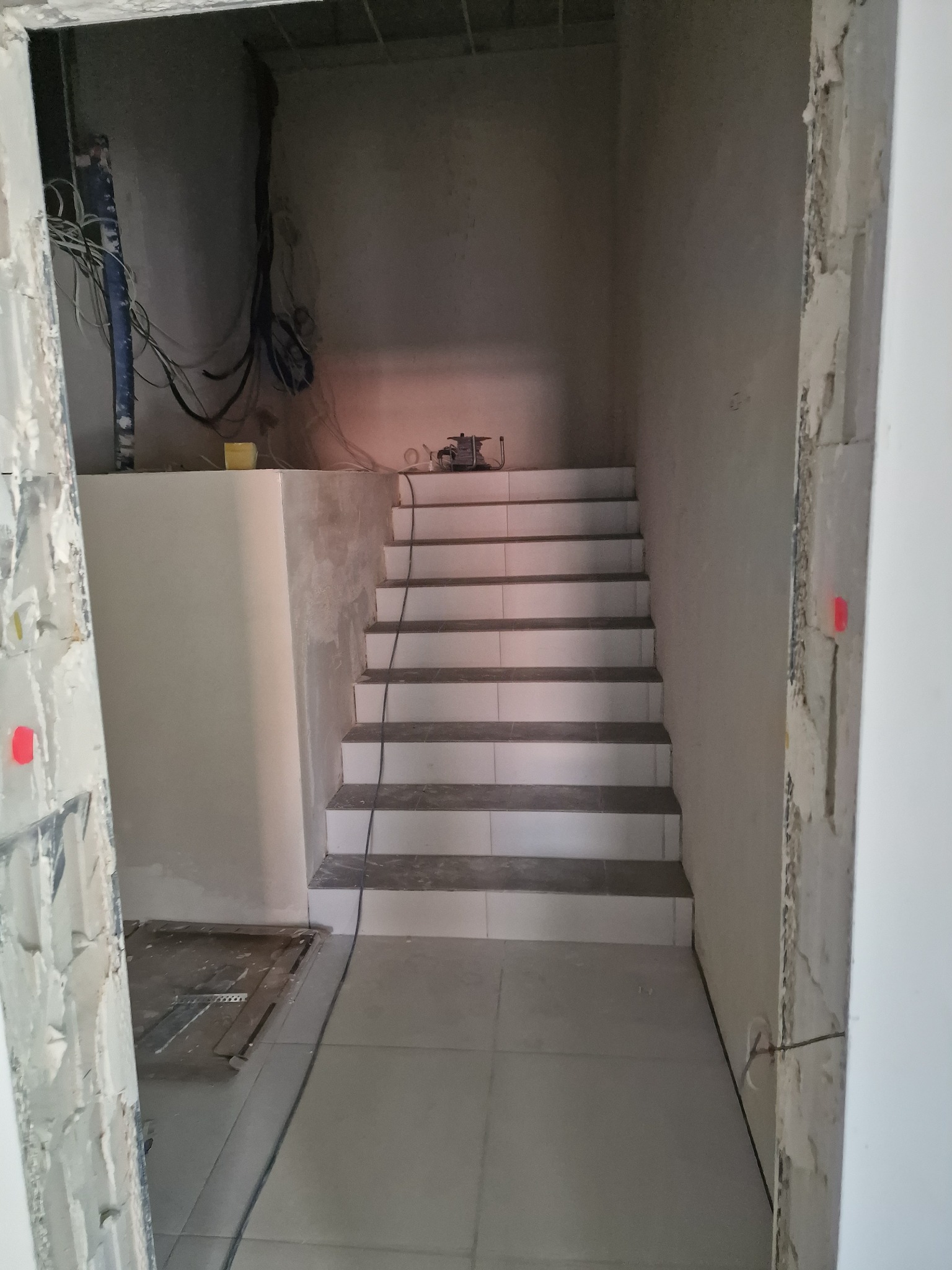 Wyłożone płytkami schody w surowym stanie, widoczne kable i narzędzia na górze schodów, niedokończone wykończenie ścian.