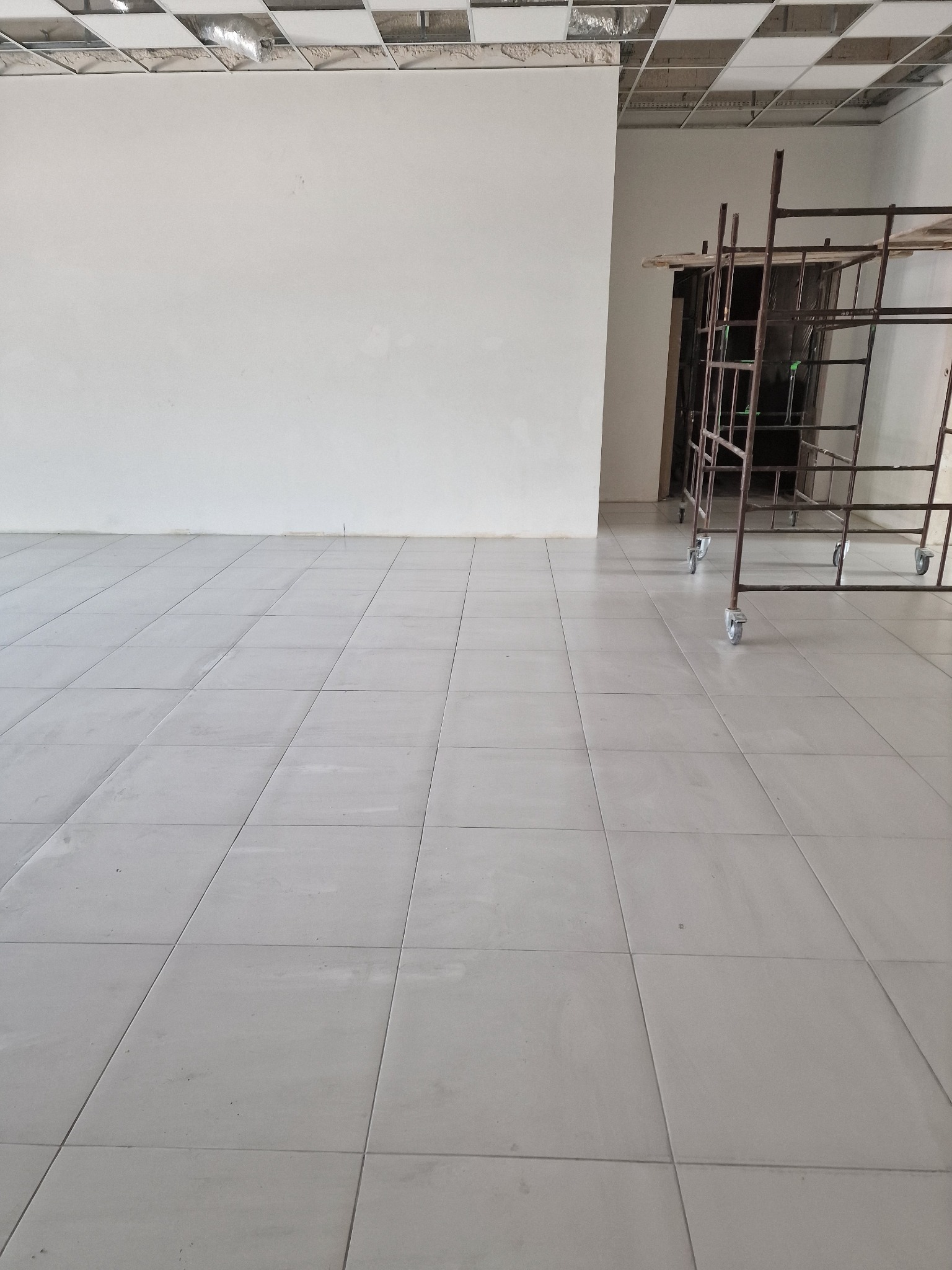 Jasne, kwadratowe płytki podłogowe w nowym pomieszczeniu z rusztowaniem na kółkach, widok na sufit z odsłoniętą instalacją.