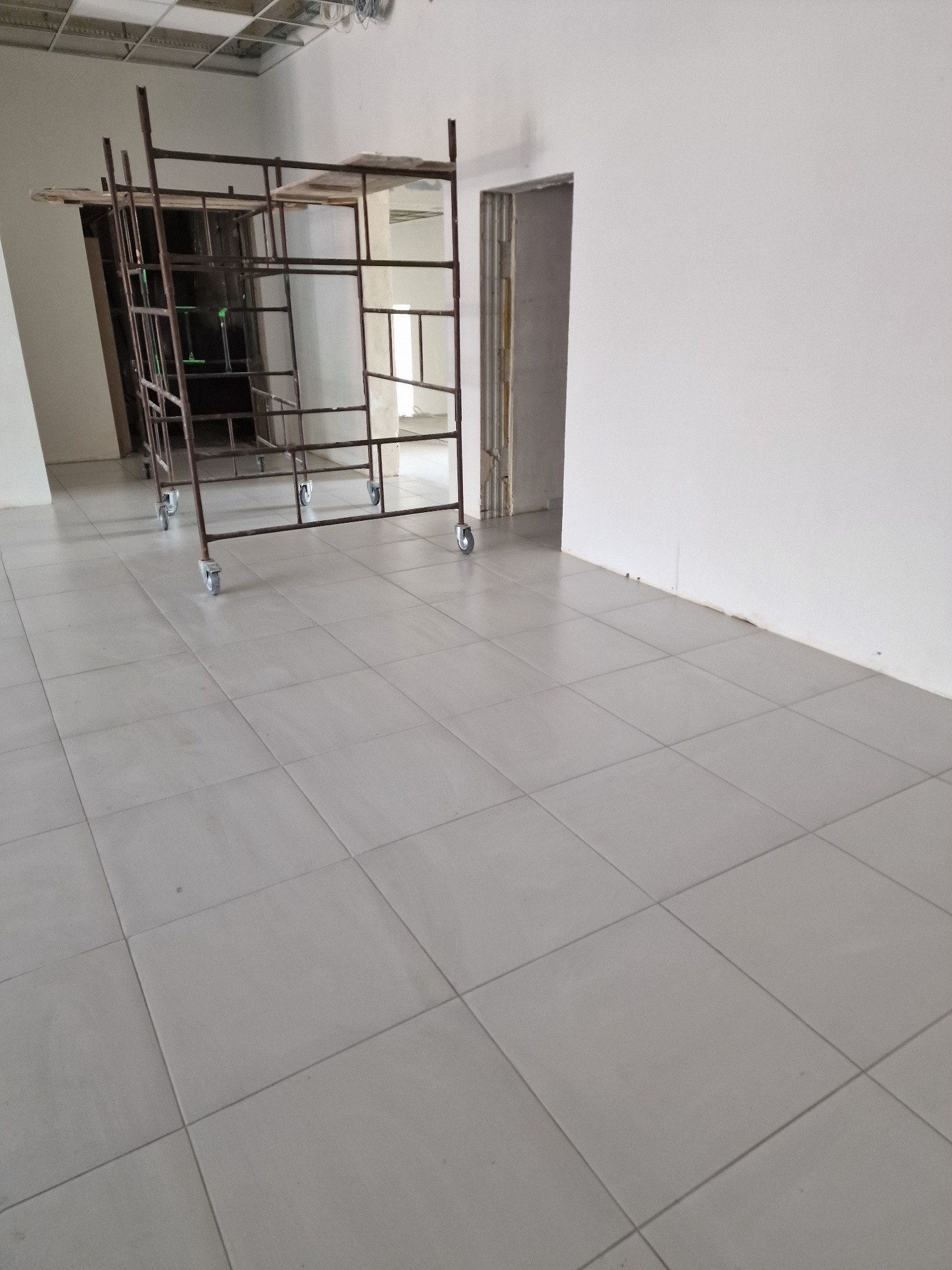 Szare płytki podłogowe ułożone w kwadraty, z metalowym rusztowaniem na kółkach w tle i fragmentem białej ściany z otworem drzwiowym.