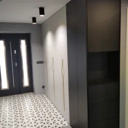 Wysoka zabudowa szafy w przedpokoju, łącząca białe fronty z minimalistycznymi złotymi uchwytami i ciemnoszary moduł z otwartą półką i szufladami, na tle wzorzystej podłogi i drzwi wejściowych.