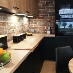 Nowoczesna kuchnia z czarnymi szafkami i drewnianymi blatami, ceglana ściana w tle, oświetlenie podszafkowe, akcesoria kuchenne na blacie.