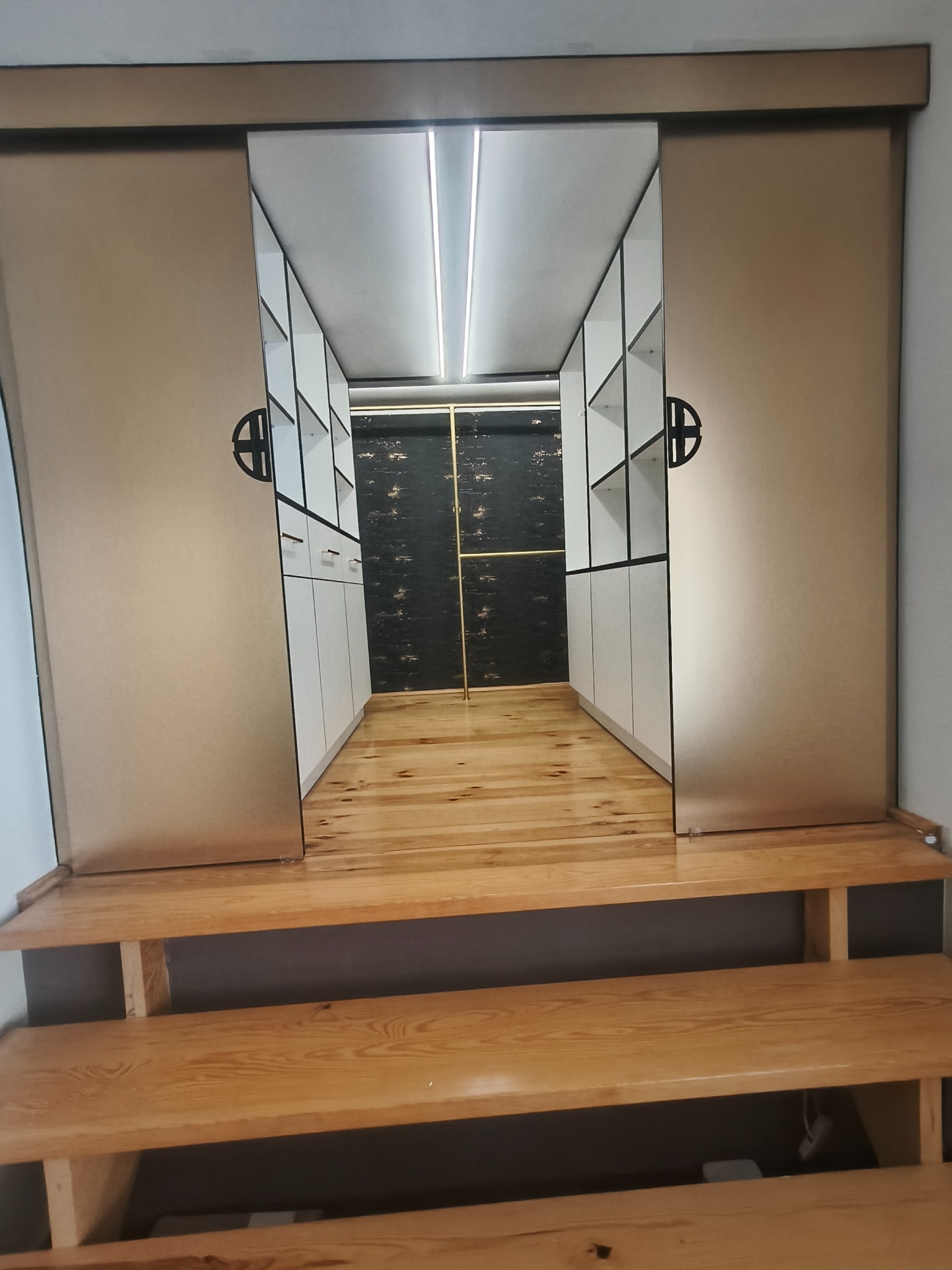 Nowoczesna garderoba z przesuwnymi drzwiami w odcieniach beżu, z drewnianą podłogą i schodami. Wnętrze szafy z białymi półkami i czarnym tłem.