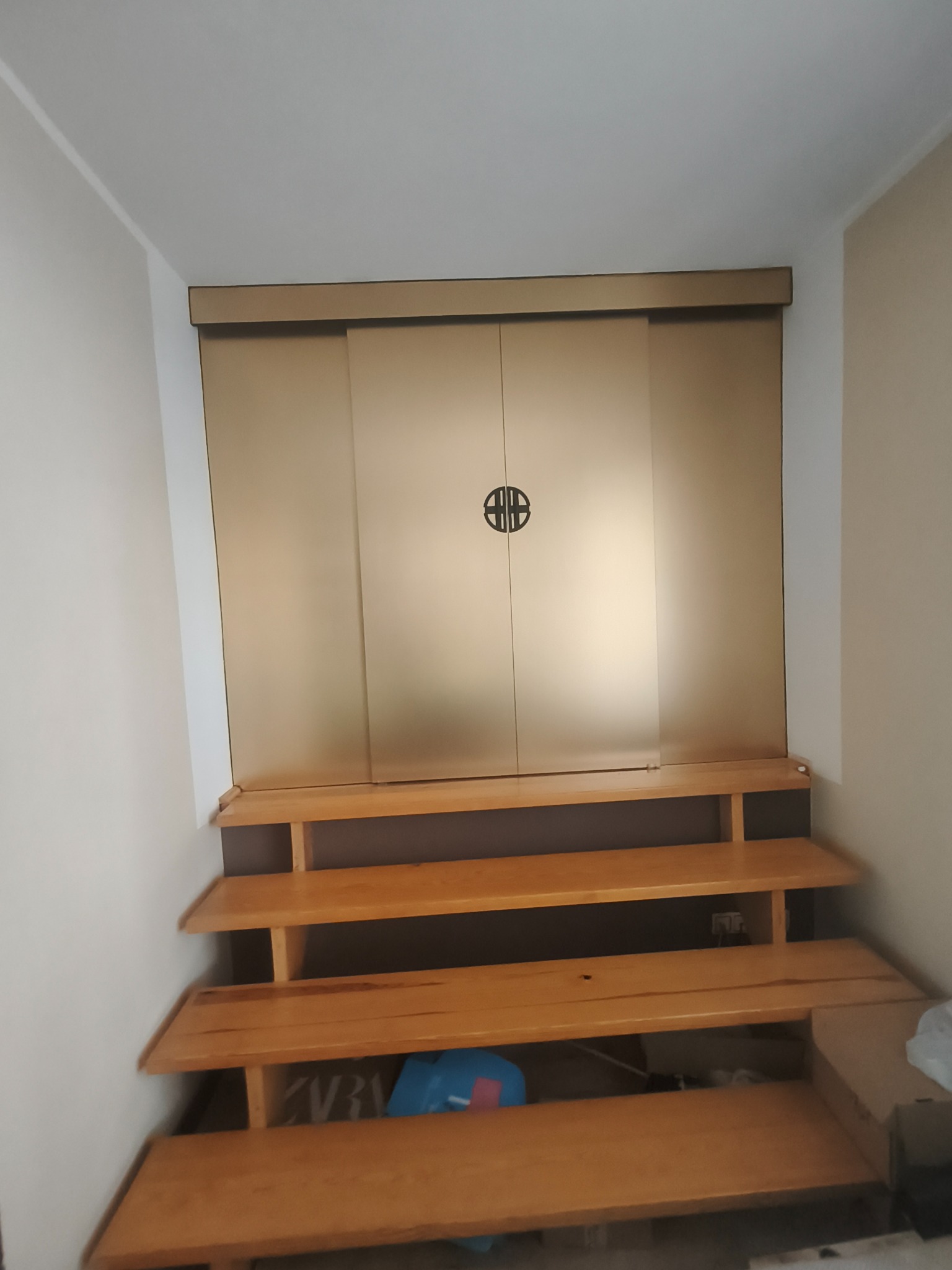 Zabudowa wnęki z przesuwnymi, złotymi drzwiami z okrągłym uchwytem, nad drewnianymi schodkowymi półkami. Widok z dołu.