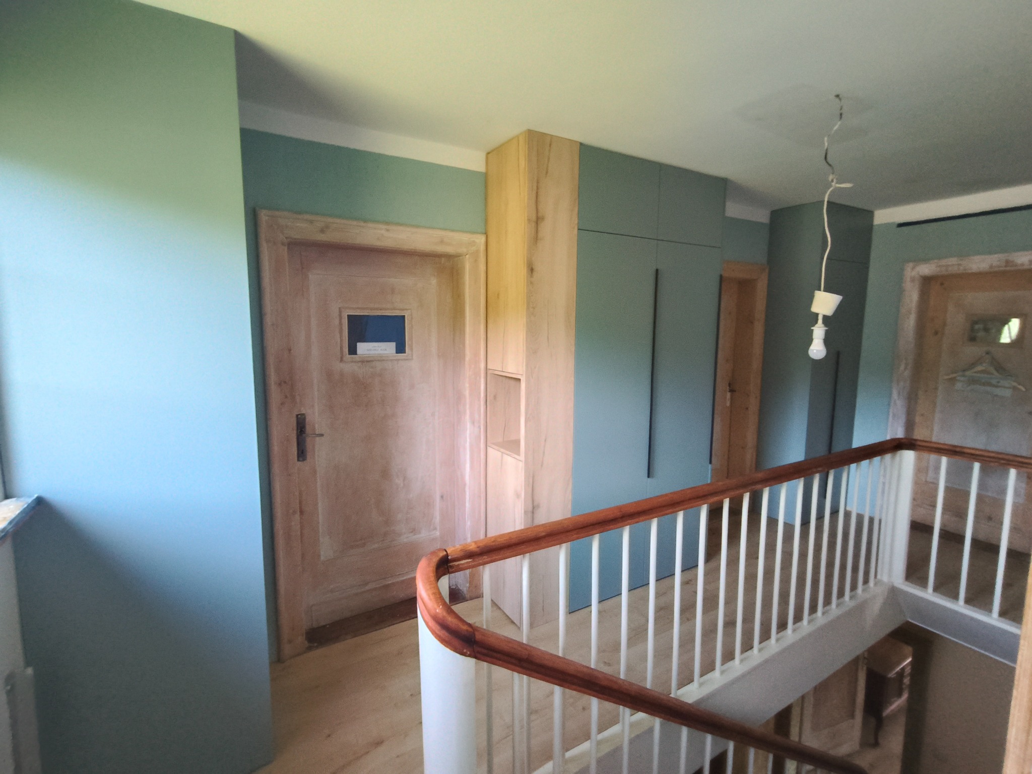 Korytarz z zabudową: szafa wnękowa i regał z jasnego drewna, drzwi z szybką, balustrada schodowa. Ściany w odcieniach zieleni i błękitu. W tle widoczne kolejne pomieszczenia.