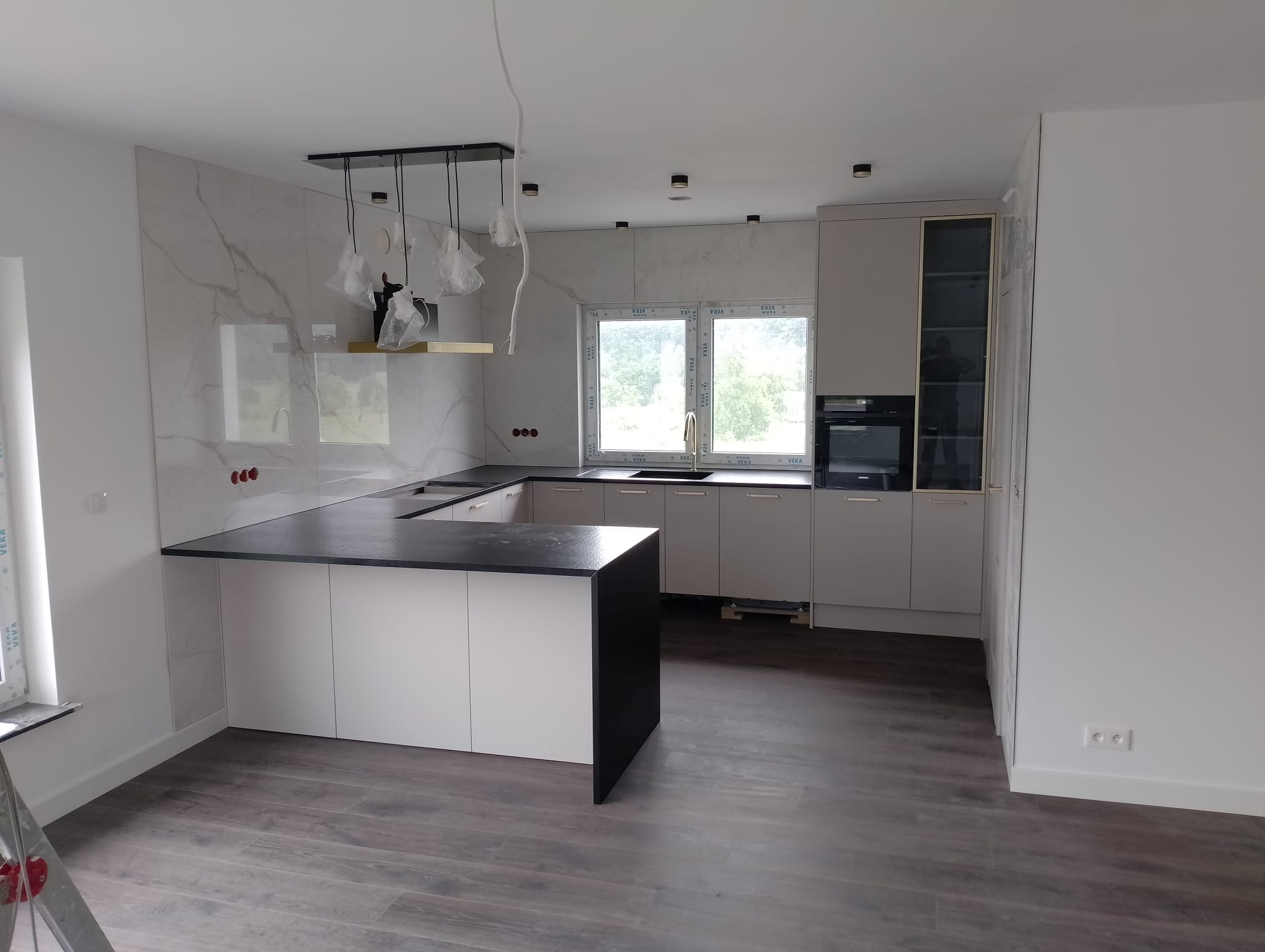 Nowoczesna kuchnia z wyspą, szarymi frontami, czarnym blatem i marmurowym backsplashiem. Widoczne okno i designerskie lampy. Podłoga z paneli imitujących drewno.