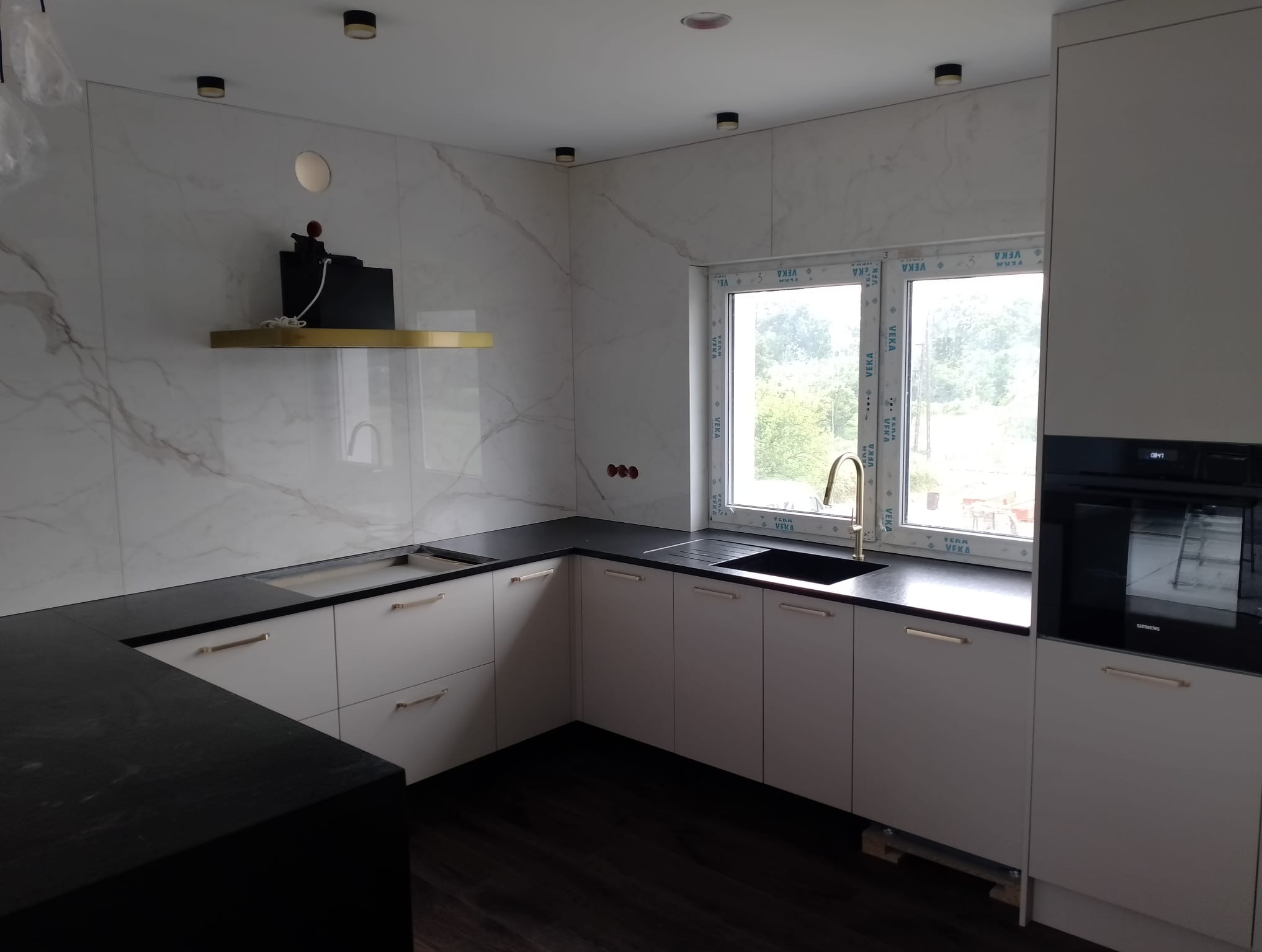 Nowoczesna kuchnia z białymi szafkami i czarnym blatem, złota bateria przy oknie. Marmurowe płytki na ścianie i czarne, drewniane panele na podłodze.