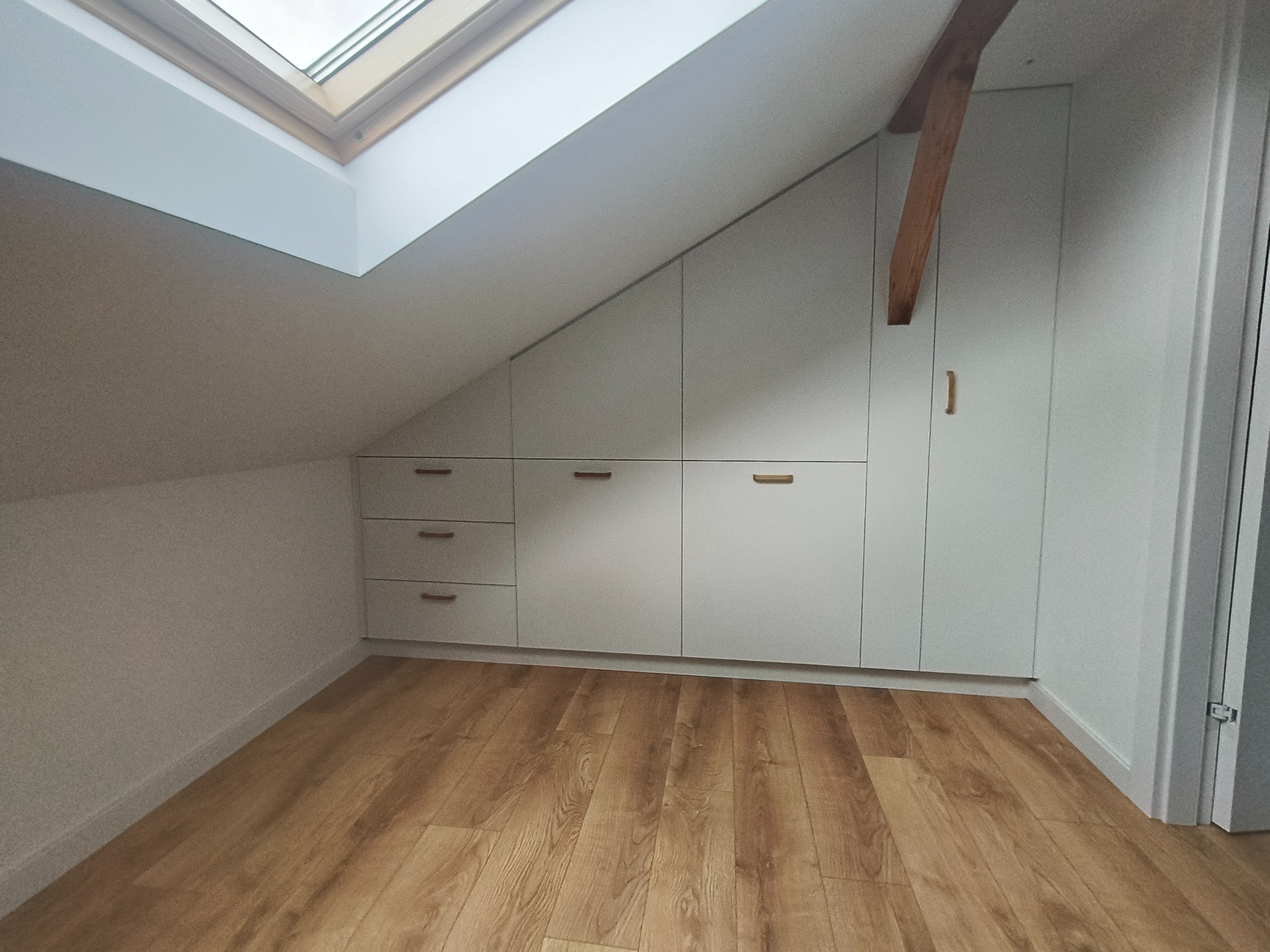 Białe szafki na wymiar, wkomponowane w skos dachu, z drewnianymi uchwytami, nad brązową podłogą. Widoczne okno dachowe i drewniana belka.