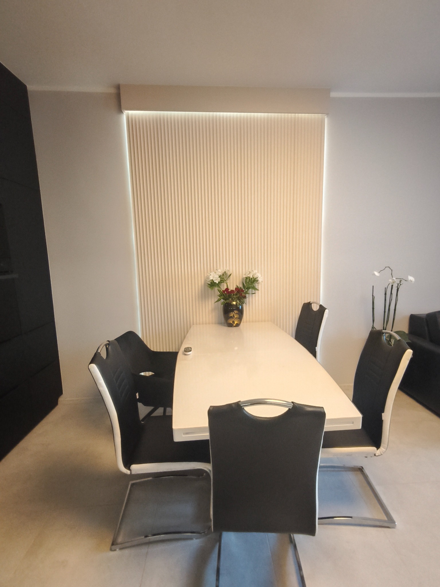 Minimalistyczna jadalnia: biały stół z kwiatami, czarno-białe krzesła na metalowych nogach, pionowe żaluzje w tle. Nowoczesne, stonowane wnętrze.