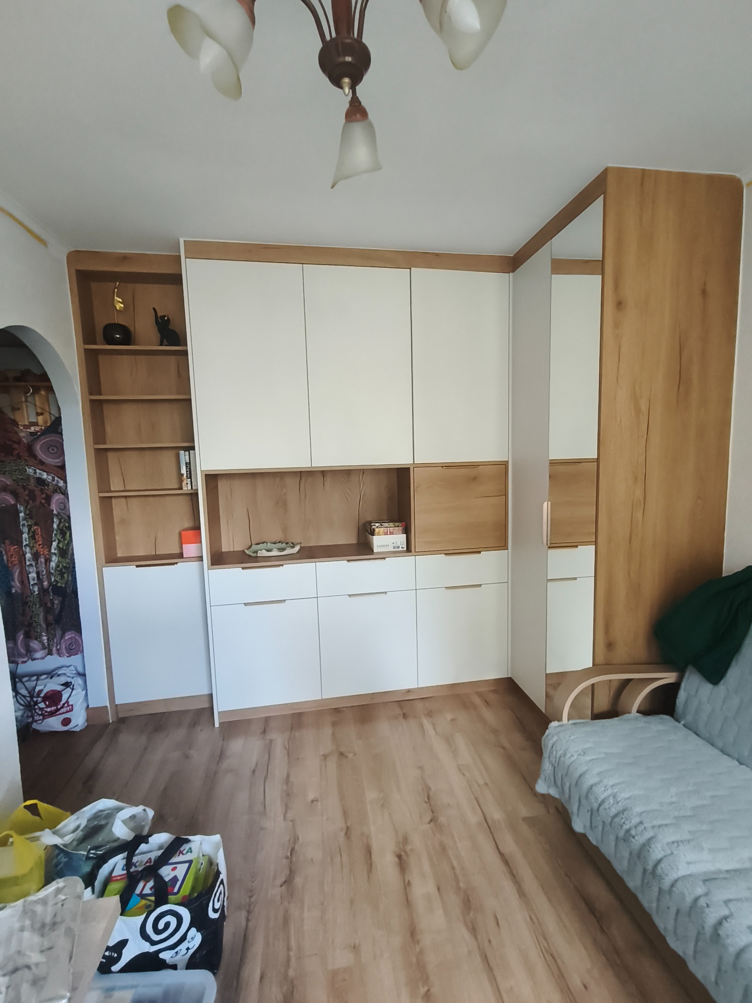 Funkcjonalna zabudowa meblowa z białymi frontami i drewnopodobnym wykończeniem, z otwartymi półkami i szufladami, uzupełniona o lustro i oświetlenie sufitowe.