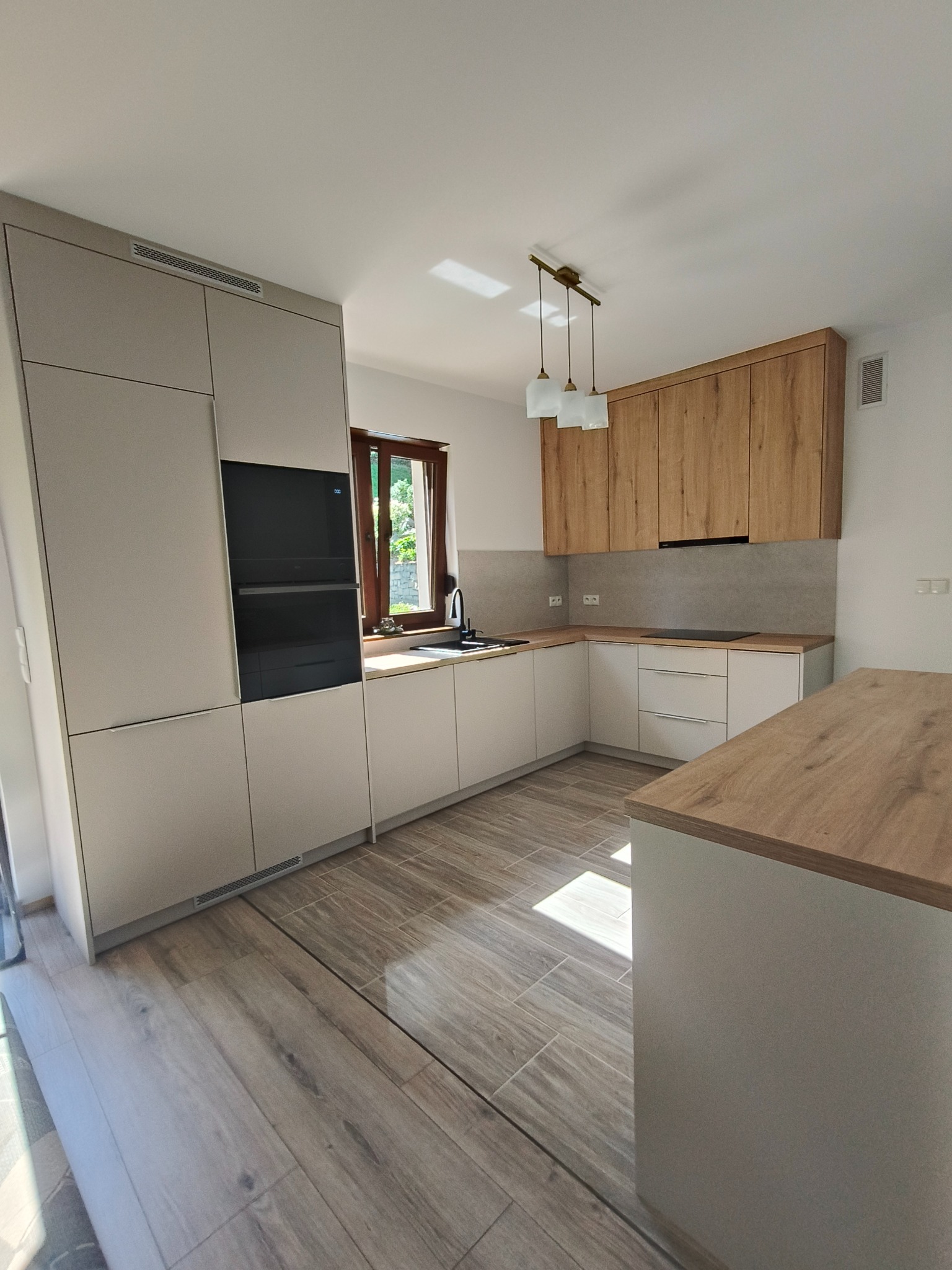 Nowoczesna kuchnia z beżowymi szafkami, drewnianymi akcentami i wyspą. Uwagę przykuwają minimalistyczne lampy wiszące i okno z widokiem na zieleń.