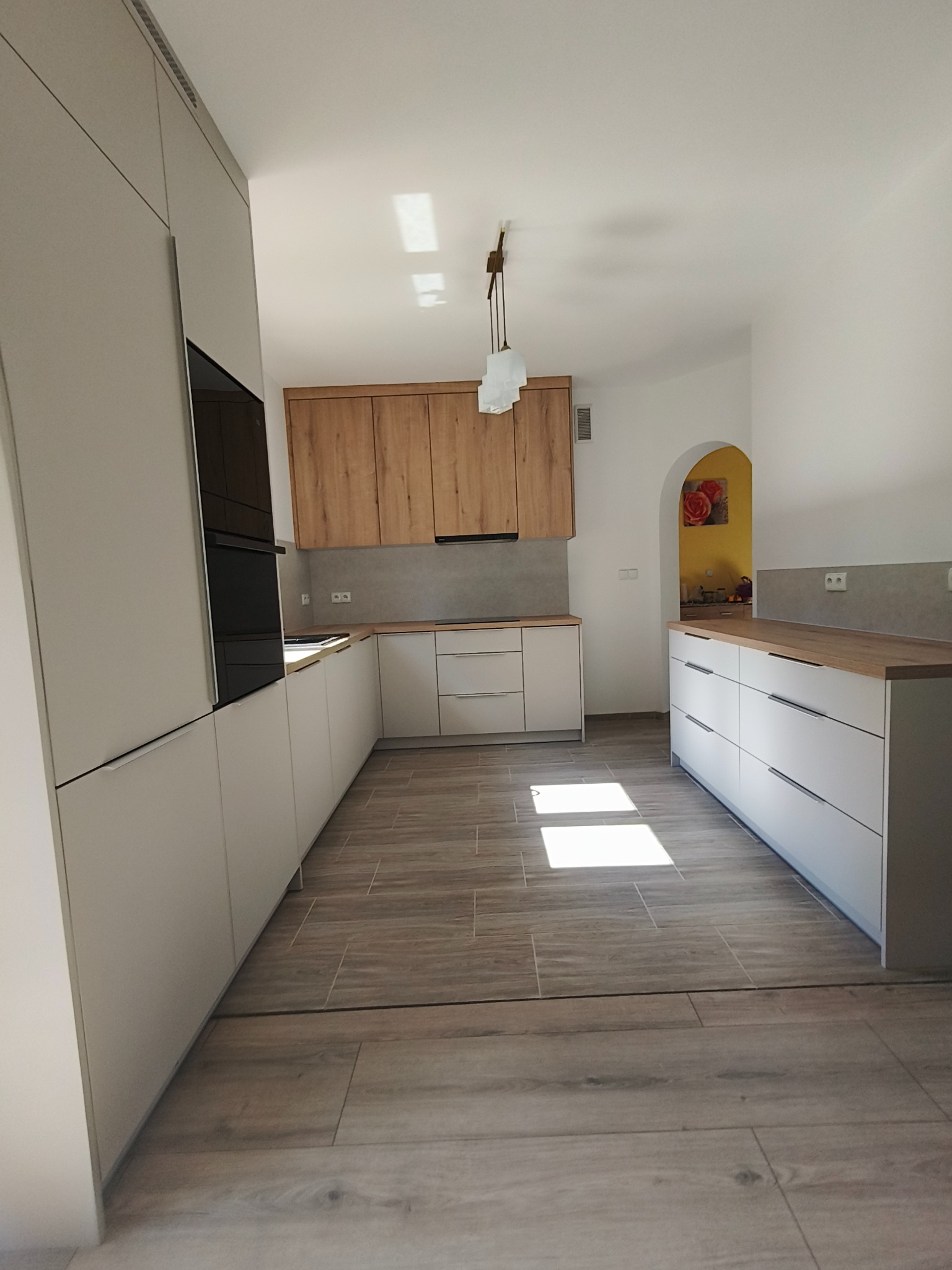 Jasna kuchnia z drewnianymi akcentami: szafki górne z naturalnego drewna, białe fronty dolne i blat. Widoczne płytki podłogowe imitujące drewno i dekoracja w przejściu.