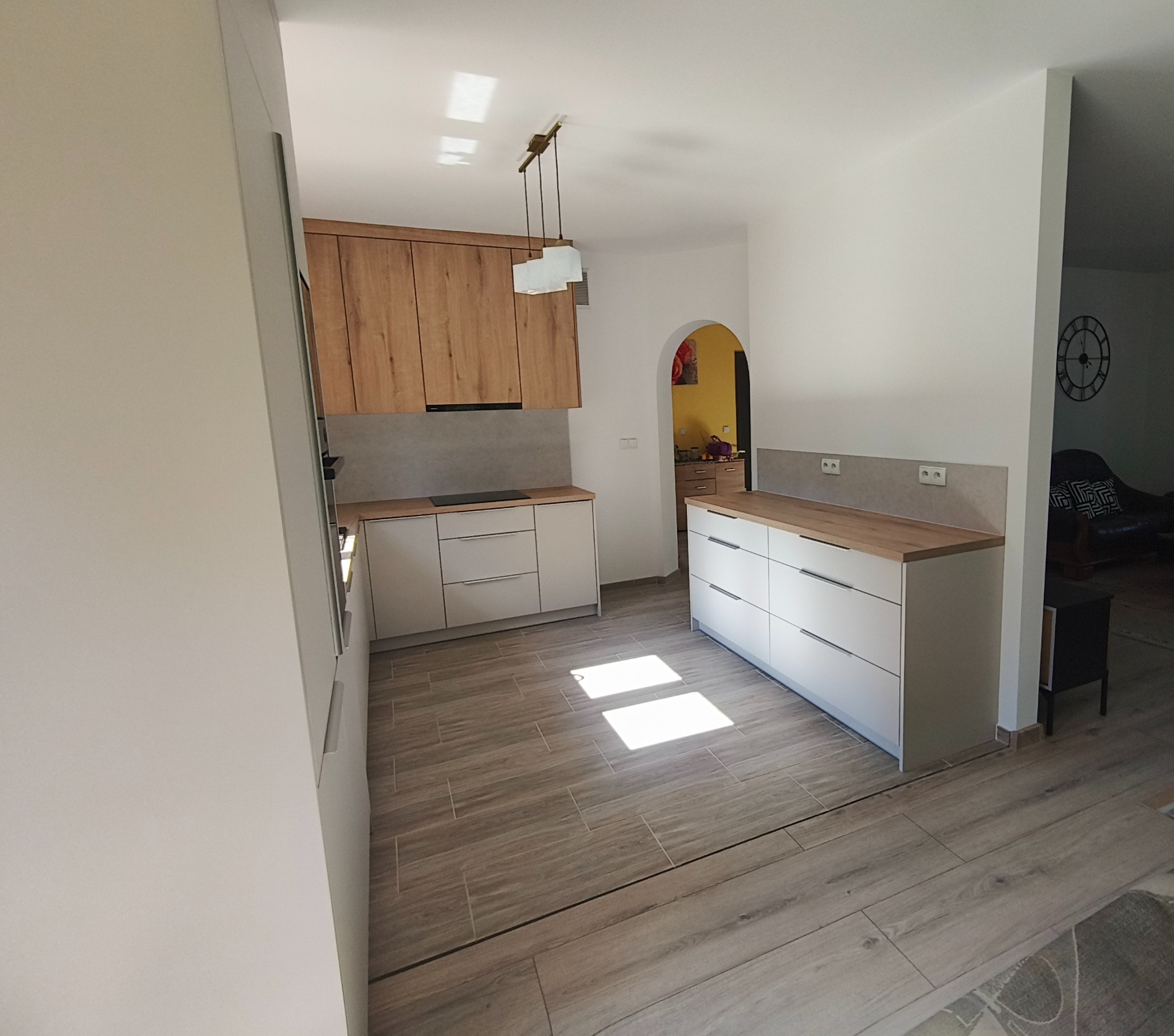 Jasna kuchnia na wymiar z drewnianymi akcentami. Widok na szafki górne z fornirem i białe, minimalistyczne szafki dolne z drewnianym blatem. Widoczne oświetlenie podszafkowe.