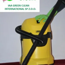 Żółty odkurzacz przemysłowy Kärcher z czarną rurą i końcówką na czerwonej wykładzinie, obsługiwany przez osobę w zielonej rękawicy, z widocznym logo firmy IAA Green Clean w tle.