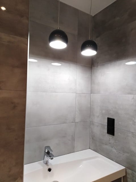 Nowoczesna łazienka z szarymi płytkami imitującymi beton, umywalką nablatową i dwoma okrągłymi lampami wiszącymi z czarnymi kloszami, oświetlona światłem punktowym.