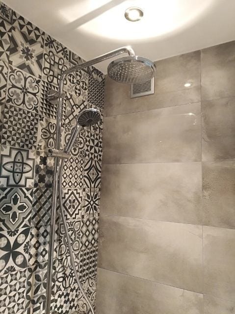 Nowoczesna kabina prysznicowa z dwoma słuchawkami i deszczownicą, wykończona płytkami w geometryczne wzory oraz szarymi, gładkimi płytami imitującymi beton.