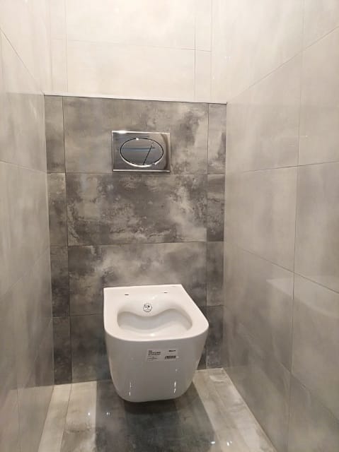 Nowoczesna, podwieszana toaleta w łazience wykończonej szarymi płytkami w Łodzi. Widoczny chromowany przycisk spłuczki.