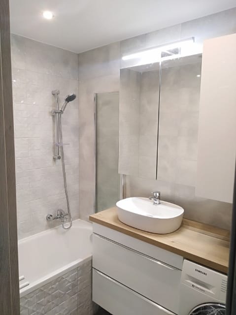 Nowoczesna łazienka po remoncie: wanna z prysznicem, umywalka nablatowa na drewnianym blacie, białe szafki z pralką, lustro z oświetleniem LED i szare płytki na ścianach.