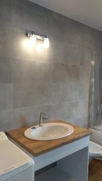 Umywalka owalna nablatowa na drewnianym blacie w łazience z szarymi płytkami, oświetlona lampą z dwoma żarówkami.