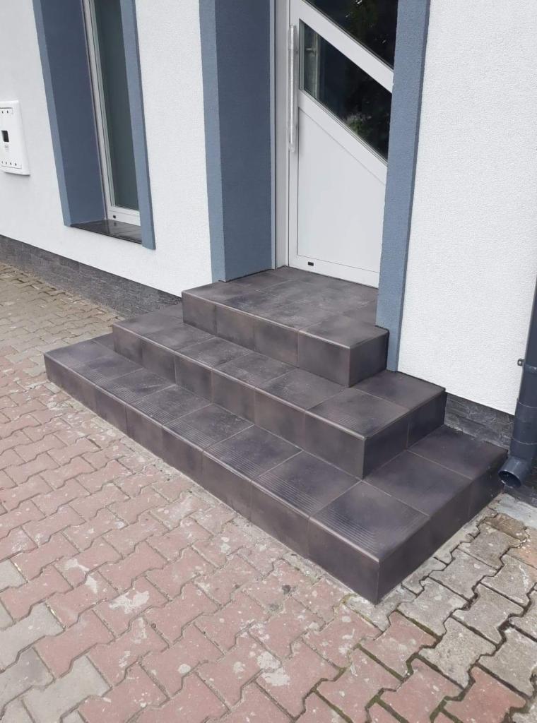 Zewnętrzne schody wejściowe wyłożone ciemnobrązowymi płytkami ceramicznymi z delikatnym wzorem, prowadzące do białych drzwi z przeszklonymi elementami i szarymi obramowaniami, na tle białej ściany...