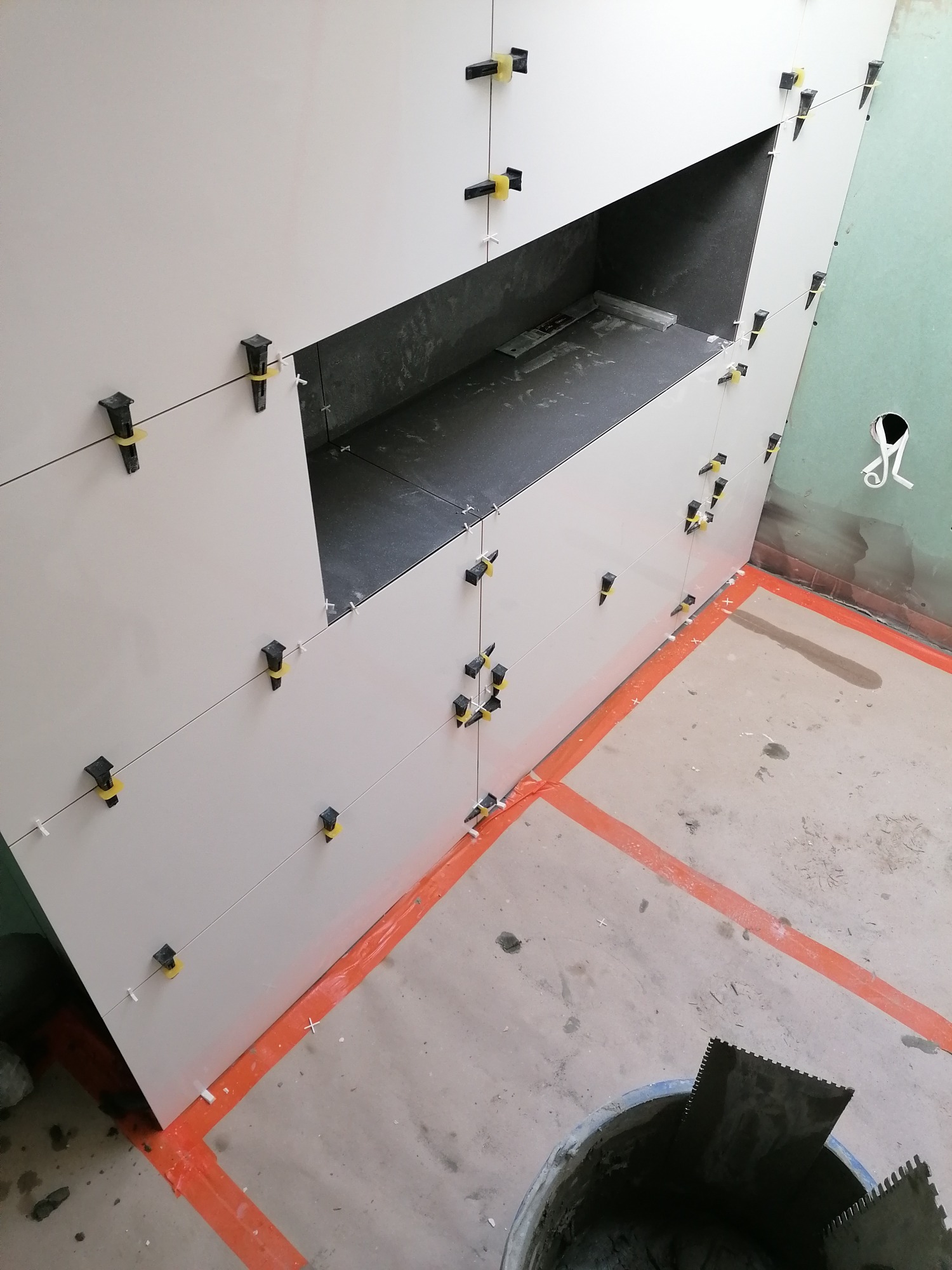 Świeżo ułożone, jasne płytki na ścianie z wnęką, zabezpieczone czarnymi klipsami poziomującymi z żółtymi gumkami, widoczne narzędzia glazurnicze i wiadro z zaprawą na podłodze zabezpieczonej...