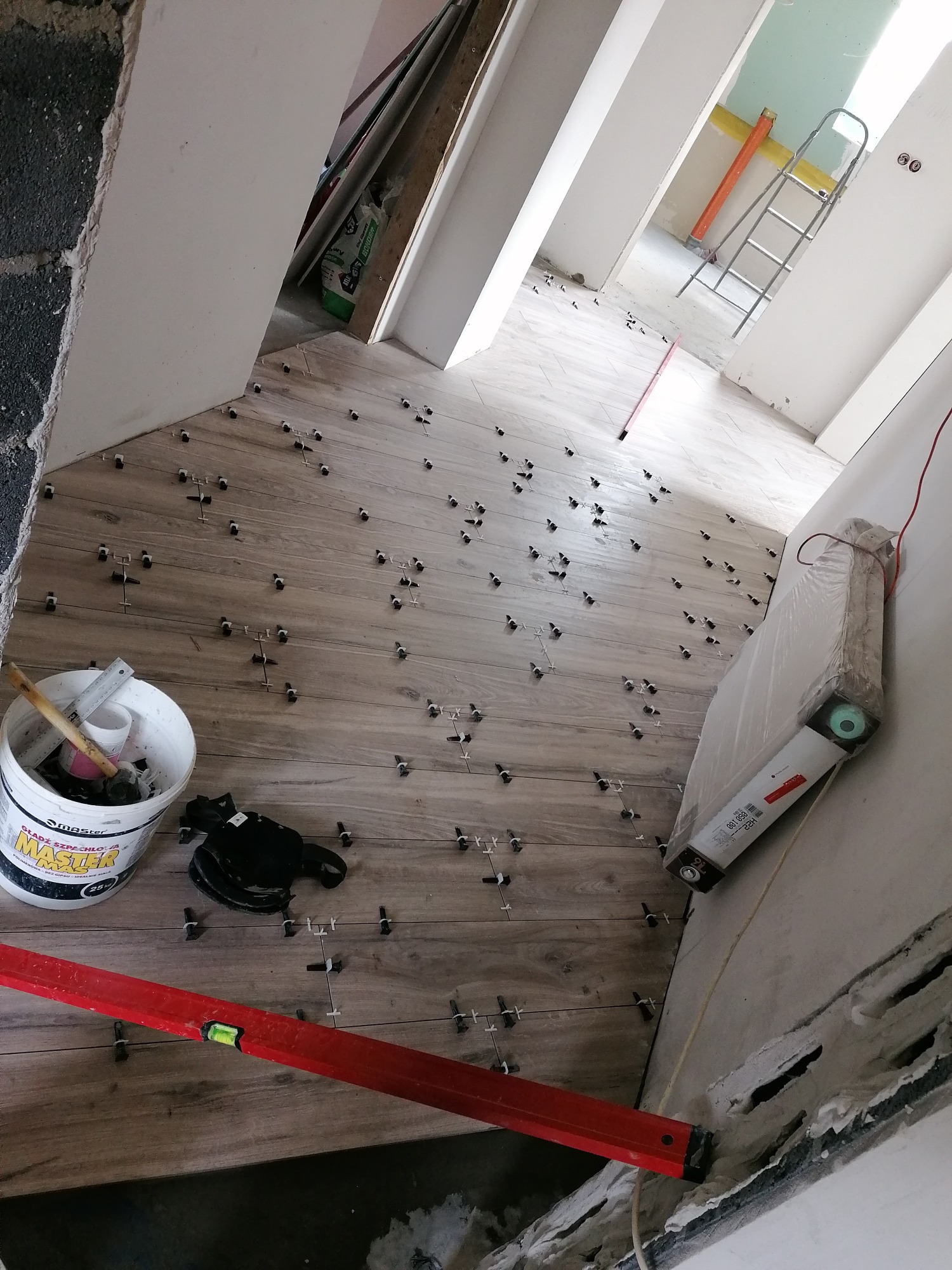 Układanie płytek podłogowych w trakcie pracy, widoczne krzyżyki dystansowe, poziomica, wiadro z zaprawą i drabina w tle.