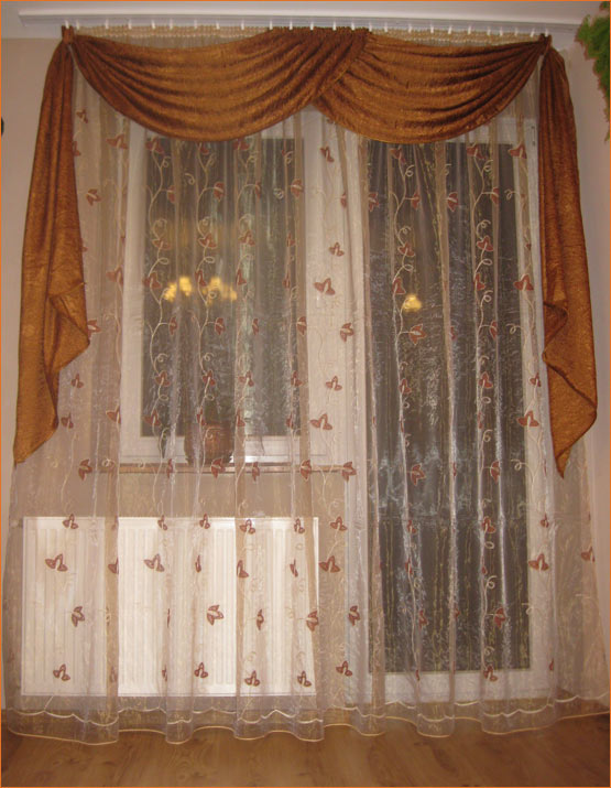 Zasłony z woalu z motywem liści i brązowym drapowaniem nad oknem, widoczny grzejnik pod oknem.