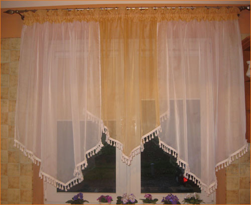 Okno udekorowane asymetrycznymi, białymi i złotymi firanami z dekoracyjnymi frędzlami, nad oknem fioletowe fiołki w doniczkach.