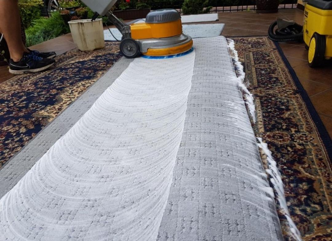 Fragment dywanu czyszczonego maszyną z żółtą obudową, widoczna piana i wzór dywanu po lewej stronie przed czyszczeniem.