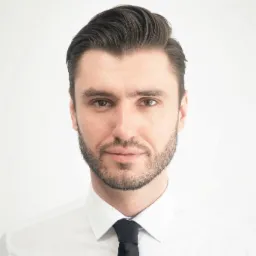 Portret biznesowy mężczyzny z ciemnymi włosami i brodą, ubrany w białą koszulę i czarny krawat, na jasnym tle. Delikatny uśmiech, profesjonalny wygląd.