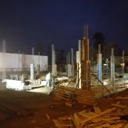 Nocny widok placu budowy z widocznymi zbrojeniami kolumn, szalunkami i pracownikami w odblaskowych kamizelkach, oświetlonego sztucznym światłem.