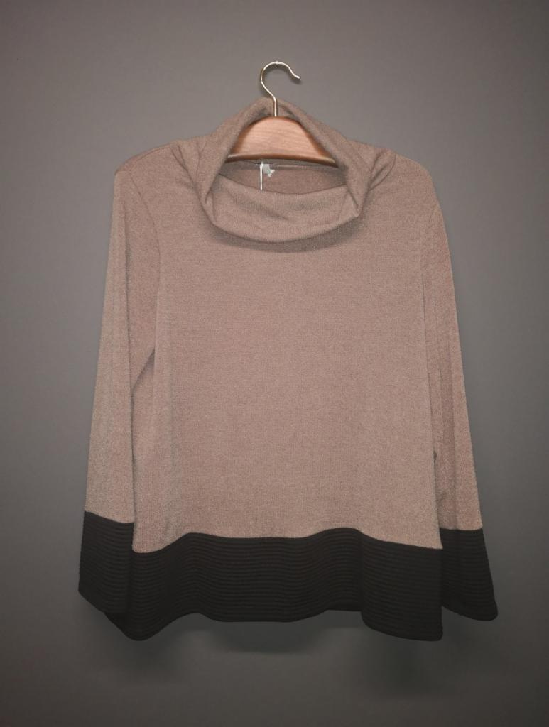 Beżowy sweter z golfem i czarnym prążkowanym dołem zawieszony na wieszaku na tle szarej ściany.