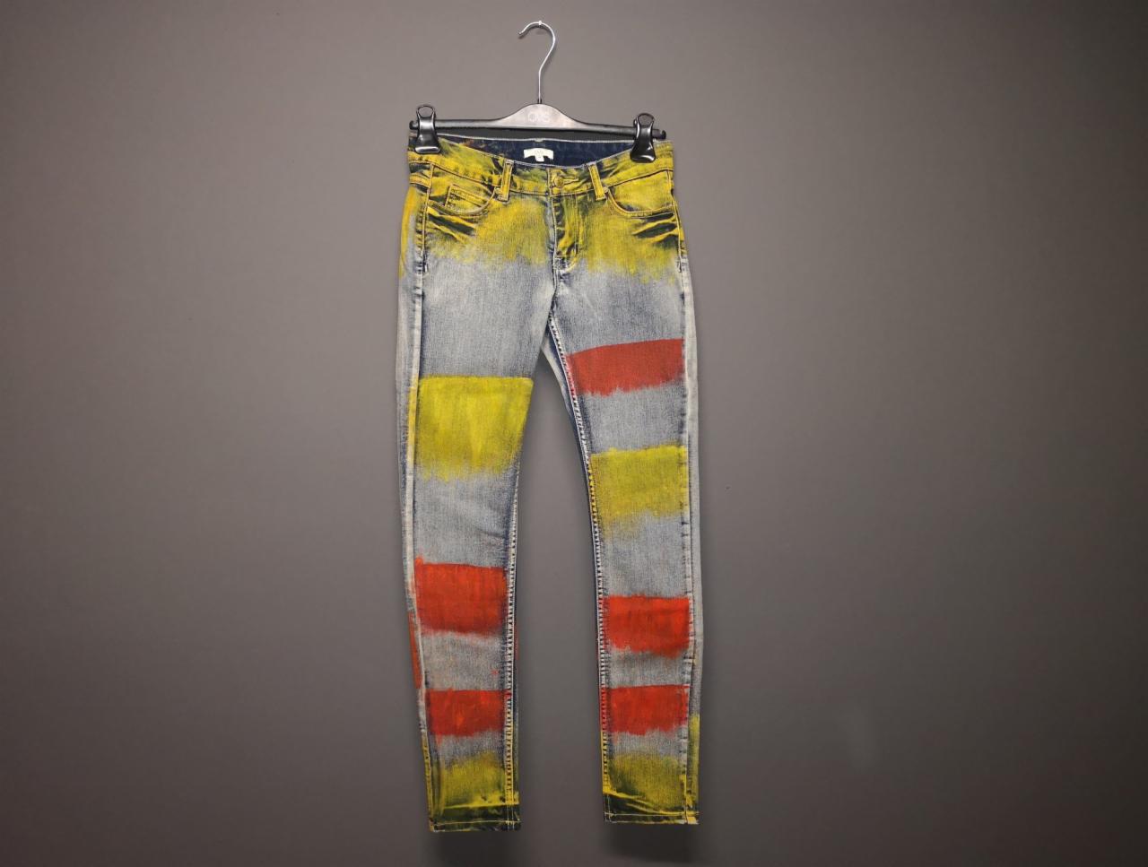 Dżinsowe spodnie typu skinny z abstrakcyjnymi, poziomymi pasami w kolorach żółtym i czerwonym, wiszące na wieszaku na tle szarej ściany.