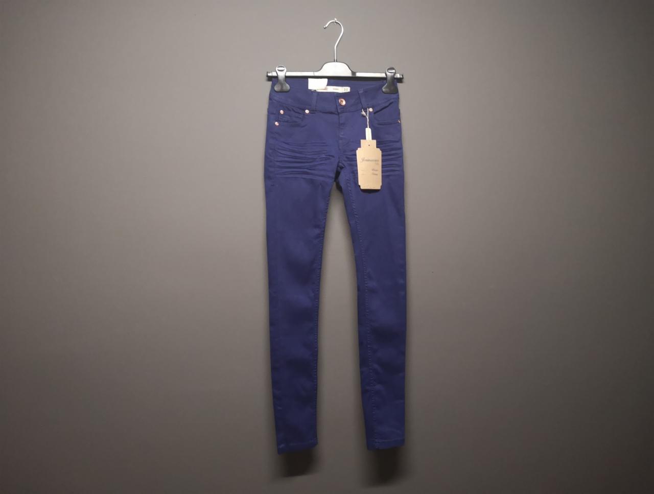 Granatowe, dopasowane spodnie damskie typu skinny jeans, wiszące na wieszaku na tle szarej ściany, z widoczną papierową metką.