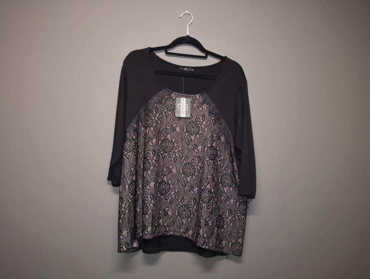 Czarna bluzka z długim rękawem i koronkowym panelem z przodu, wisząca na czarnym wieszaku na tle szarej ściany, widoczna metka.