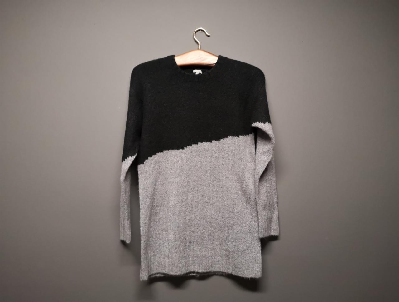 Damski sweter oversize z grubego, melanżowego materiału w kolorach czarnym i szarym, wiszący na drewnianym wieszaku na tle szarej ściany.