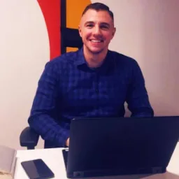 Uśmiechnięty mężczyzna w niebieskiej koszuli w kratę siedzi przy biurku z laptopem i telefonem, w tle abstrakcyjna grafika ścienna w kolorach czerwonym, żółtym, zielonym i białym.