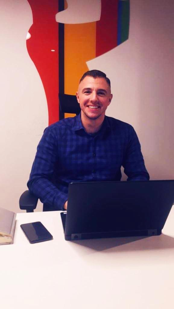 Uśmiechnięty mężczyzna w niebieskiej koszuli w kratę siedzi przy biurku z laptopem i telefonem, w tle abstrakcyjna grafika ścienna w kolorach czerwonym, żółtym, zielonym i białym.