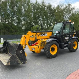 Żółto-czarna ładowarka teleskopowa JCB z łyżką na kostce brukowej, widok z boku, z zielonymi drzewami w tle.