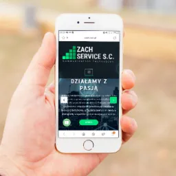 Osoba trzyma w dłoni smartfon, na którego ekranie wyświetla się strona internetowa firmy ZACH SERVICE S.C. z logotypem i hasłem 'Działamy z pasją'.