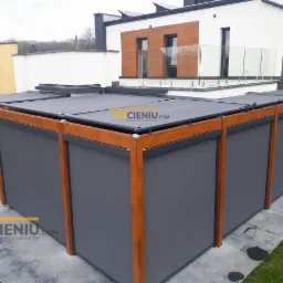 Zadaszona pergola z drewnianą ramą i szarymi, zwijanymi osłonami bocznymi, widok z zewnątrz na tle nowoczesnego domu.