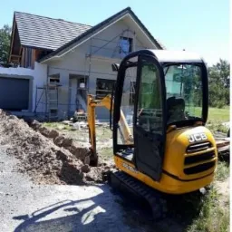 Żółta mini koparka JCB wykonuje wykop w ziemi obok domu w trakcie budowy, widoczne rusztowania i materiały budowlane.