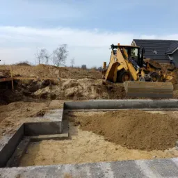 Żółta koparko-ładowarka CAT 432F przy wykopie fundamentowym z betonowym szalunkiem, widoczny grunt i częściowo wykopana ziemia, w tle dom z ciemnym dachem i błękitne niebo.