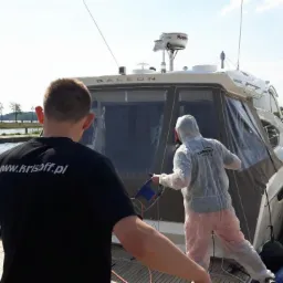 Czyszczenie jachtu Galeon przez dwóch mężczyzn na pomoście. Jeden w czarnej koszulce z adresem www, drugi w kombinezonie ochronnym używa elektronarzędzia.