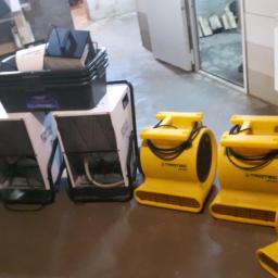 Krisoff - Rząd białych osuszaczy kondensacyjnych z czarnymi kratkami wentylacyjnymi i żółte dmuchawy marki Trotec ustawione na betonowej podłodze w pomieszczeniu z grzejnikiem na ścianie.