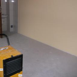 Krisoff - Żółty, przemysłowy osuszacz powietrza z czarną kratką wentylacyjną i uchwytem, ustawiony w rogu pustego, szarego pokoju z beżową ścianą i widocznym wejściem do łazienki z białą umywalką.