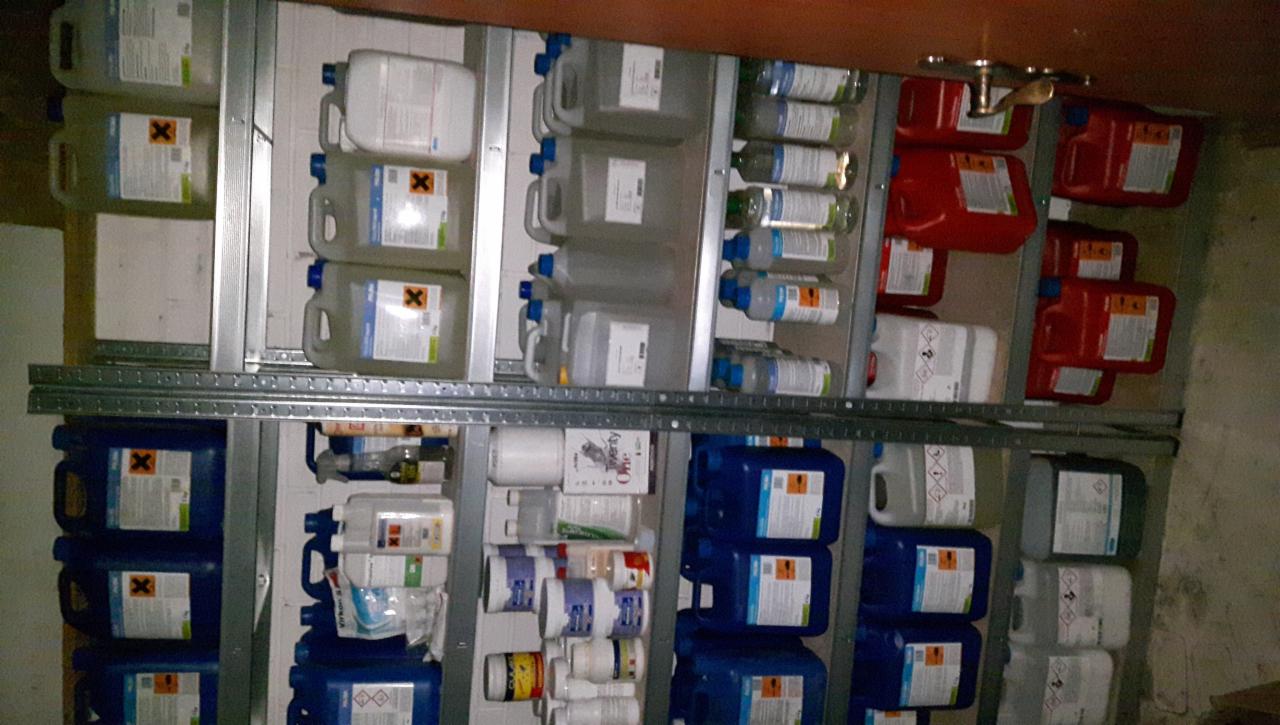 Półki magazynowe wypełnione różnokolorowymi pojemnikami z substancjami chemicznymi, niektóre oznaczone symbolem ostrzegawczym w postaci czarnego krzyża na pomarańczowym tle.