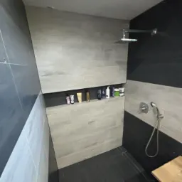 Nowoczesna kabina prysznicowa z szarymi płytkami imitującymi drewno, wnęka na kosmetyki z oświetleniem punktowym i czarnymi akcentami na ścianach.