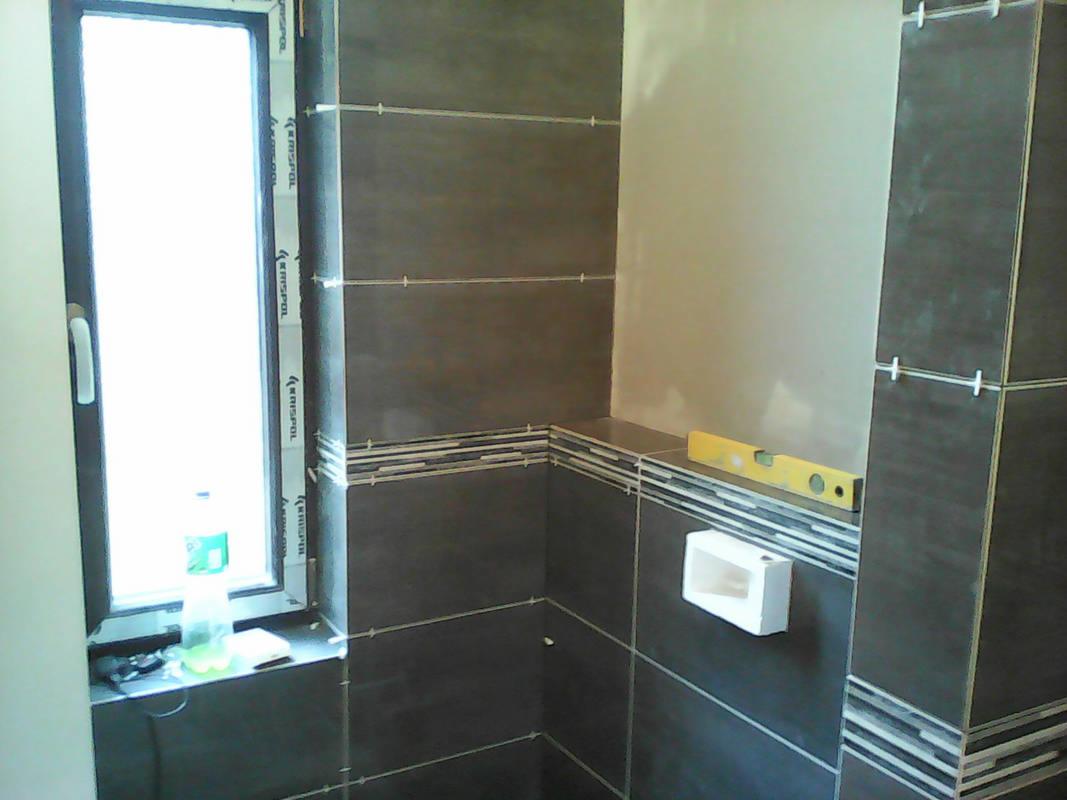 Ściana łazienki w trakcie układania ciemnych płytek, z widocznymi krzyżykami dystansowymi i poziomnicą, z fragmentem okna po lewej stronie.