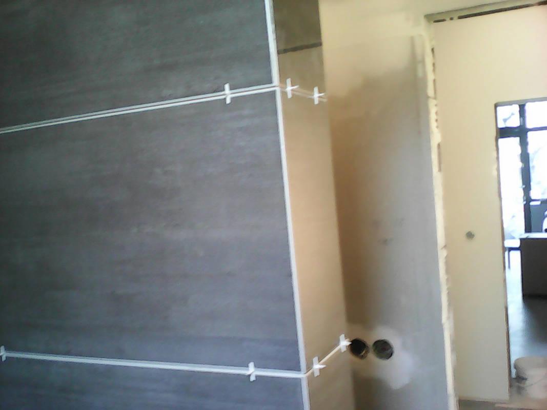 Montaż szarych płytek wielkoformatowych na ścianie z użyciem krzyżyków dystansowych, widoczne otwory pod instalacje.