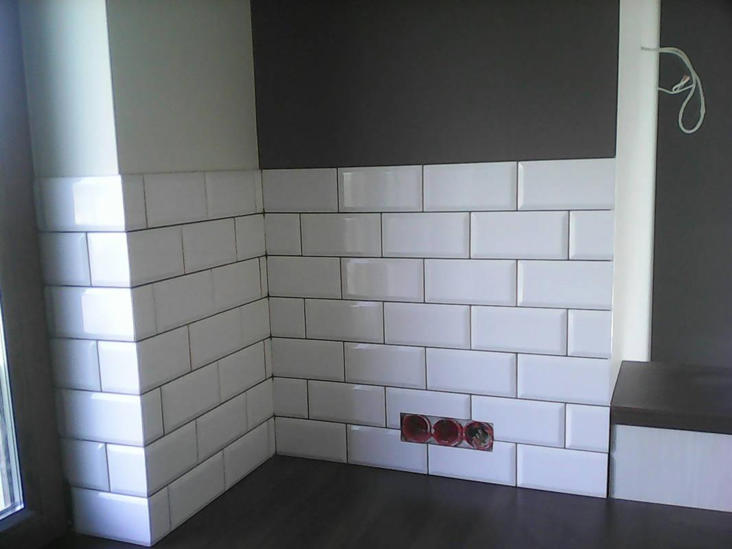 Białe, prostokątne płytki ułożone na ścianie, z widocznymi gniazdkami elektrycznymi bez osprzętu, ciemna fuga, szara ściana powyżej płytek.